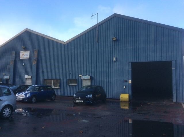 Large blue corrugated metal industrial warehouse building with multiple roller shutter doors and a car park, representative of commercial industrial asbestos survey sites.